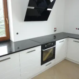 Kuchnia z czarnym blatem, białymi szafkami z falistym wzorem i czarnym okapem, widok na okno z drewnianą ramą.