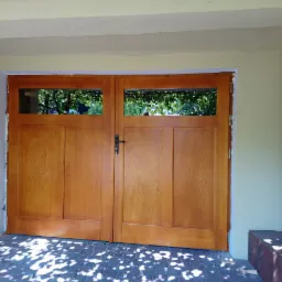 Dwu-skrzydłowe, jasne, drewniane drzwi z przeszkleniami u góry, zamontowane w jasnym budynku, widoczne cienie liści na brukowanym podłożu.