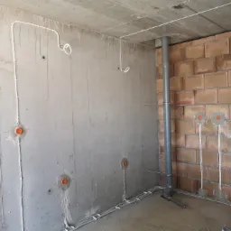 Surowy stan: Instalacja elektryczna w trakcie montażu, przewody prowadzone po ścianach z betonu i cegły, widoczne puszki instalacyjne w kolorze pomarańczowym.