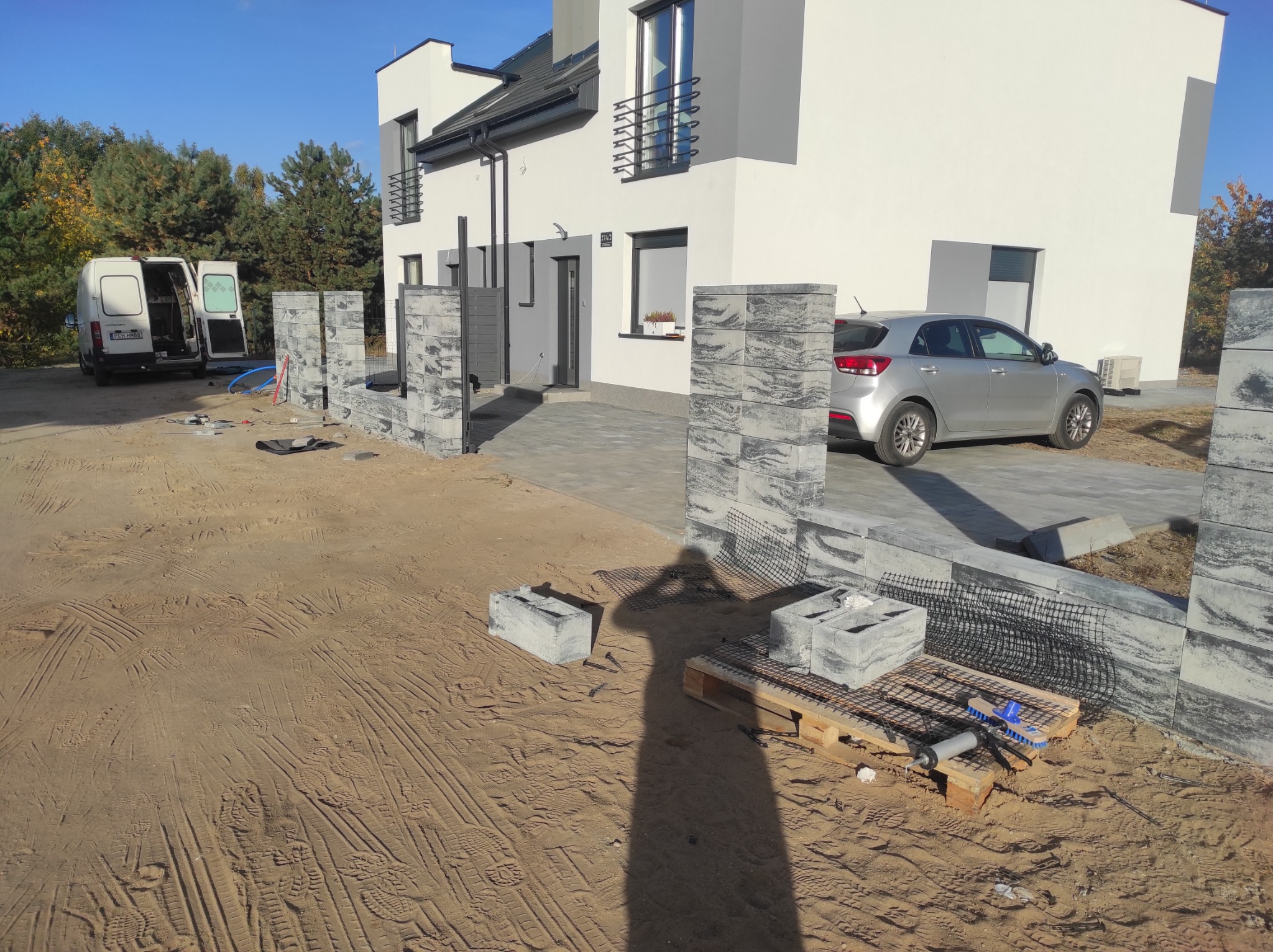 Budowa ogrodzenia z bloczków betonowych na piaszczystym terenie przed nowoczesnym domem, widoczny samochód i bus ekipy budowlanej.
