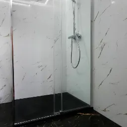 Nowoczesna kabina prysznicowa z przesuwnymi drzwiami, chromowanymi profilami i czarnym brodzikiem, na tle białych płytek imitujących marmur z delikatnym złotym żyłowaniem, oraz podłogi z czarnego...