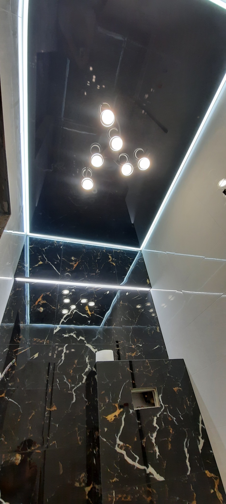 Nowoczesna łazienka z czarnymi płytkami imitującymi marmur, podświetlanym sufitem i okrągłymi lampami punktowymi, widok z dołu.