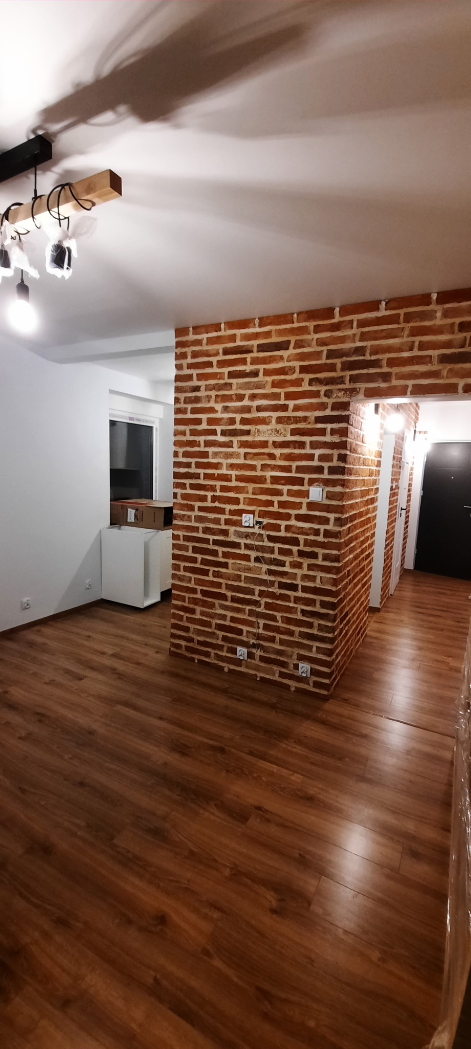 Salon po remoncie z drewnianą podłogą, ścianą z czerwonej cegły i nowoczesnym oświetleniem sufitowym. Część mebli jeszcze zabezpieczona folią.
