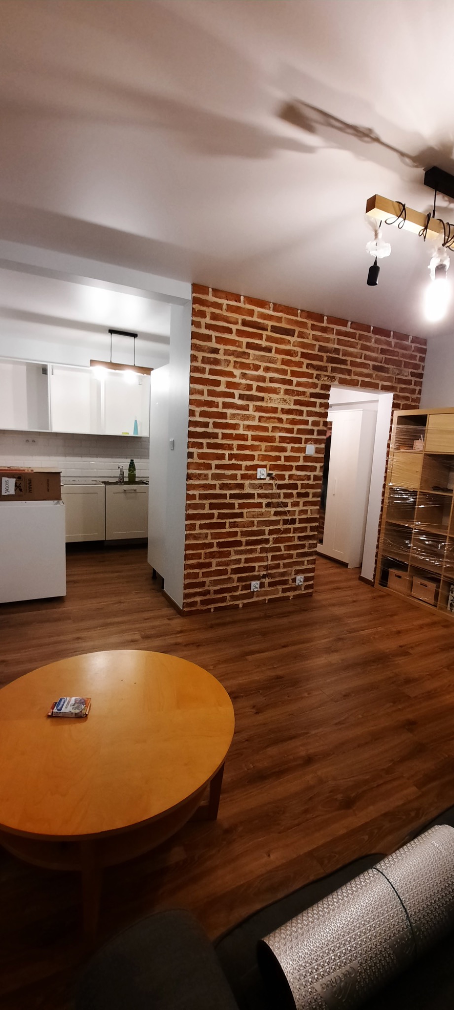 Częściowo wykończone wnętrze mieszkania z ceglaną ścianą działową, drewnianą podłogą i widoczną kuchnią w tle. Na pierwszym planie okrągły stół i zwinięta mata do ćwiczeń.