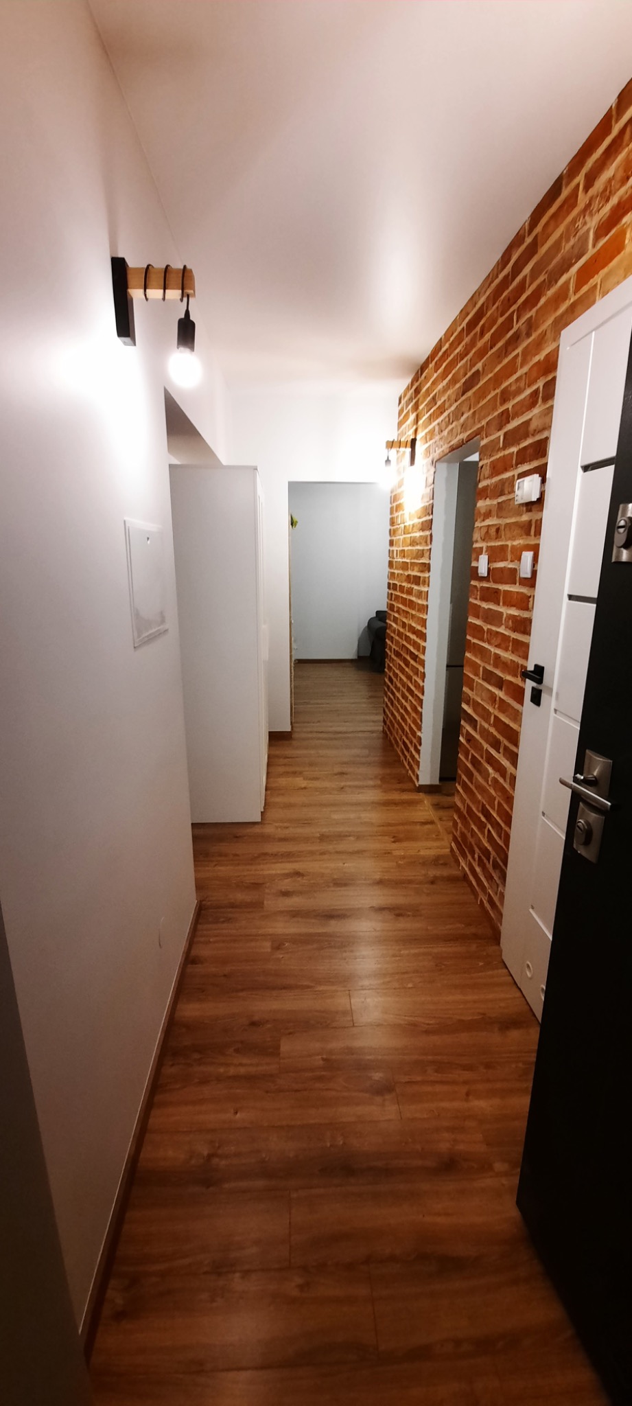 Wnętrze korytarza z panelami podłogowymi w odcieniach brązu, białą ścianą z minimalistycznym oświetleniem i ścianą z cegły, widok na otwarte przejście do kolejnego pomieszczenia.