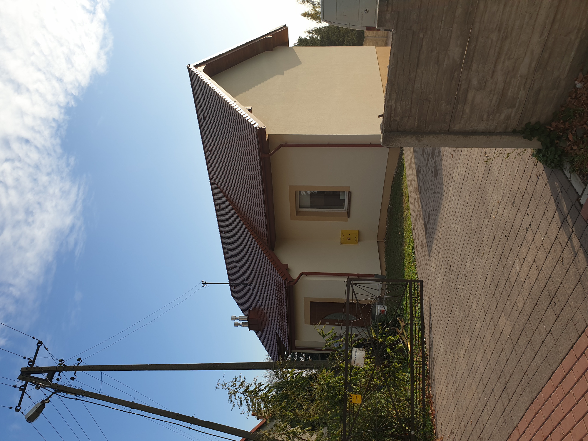 Dom jednorodzinny z beżową elewacją i czerwoną dachówką, widoczny z boku, z żółtą skrzynką pocztową i metalowym ogrodzeniem w tle.