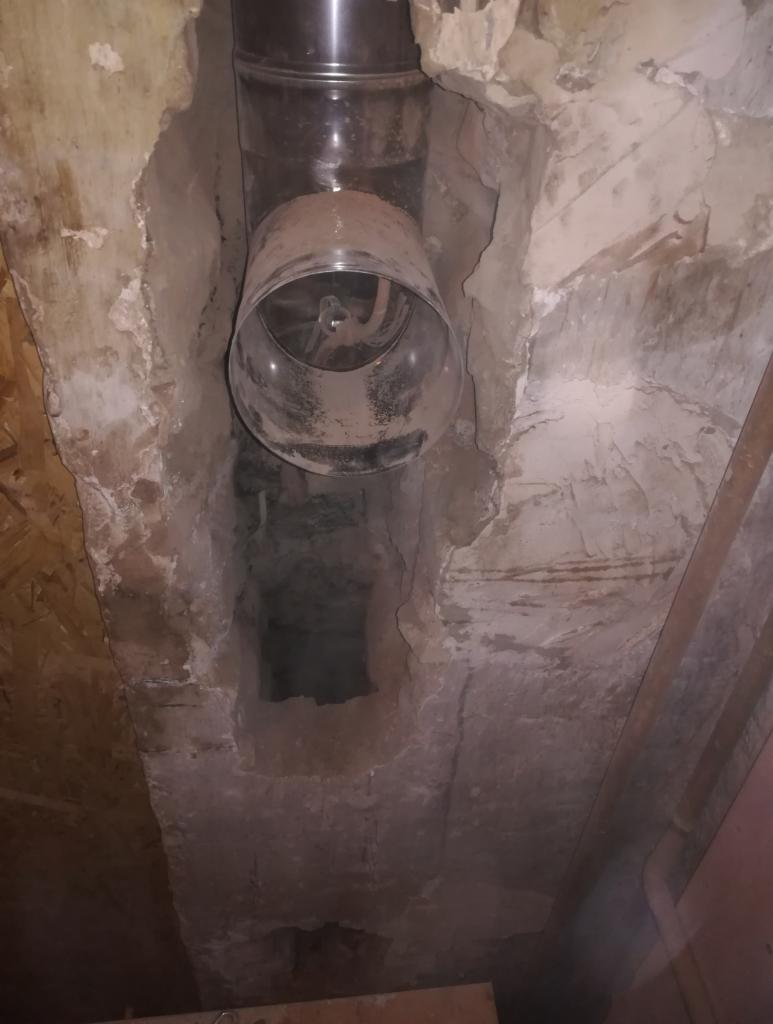 Metalowy wkład kominowy zamontowany w starym, częściowo rozkruszonym kominie murowanym, widoczne ślady sadzy i pyłu.