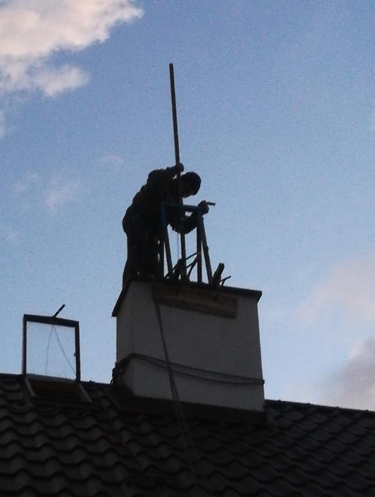 Sylwetka osoby pracującej na dachu przy kominie, widoczna na tle nieba z chmurami, z otwartym oknem dachowym w tle.