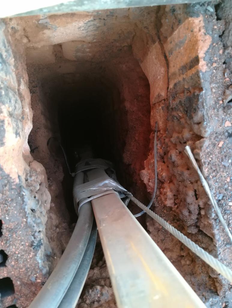 Wnętrze przewodu kominowego z cegły, widoczne przewody zabezpieczone taśmą, lina i kabel zwisające w dół.