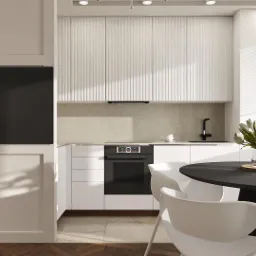 Wnętrze jasnej kuchni z białymi, ryflowanymi szafkami, okrągłym czarnym stołem jadalnym z białymi krzesłami oraz zabudowanym telewizorem w strefie wejściowej.