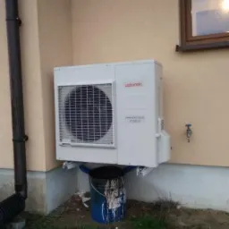 Zewnętrzna jednostka pompy ciepła marki Atlantic zamontowana na elewacji budynku, podparta nieestetycznym, pomalowanym wiadrem, widoczny kran na ścianie i rynna.