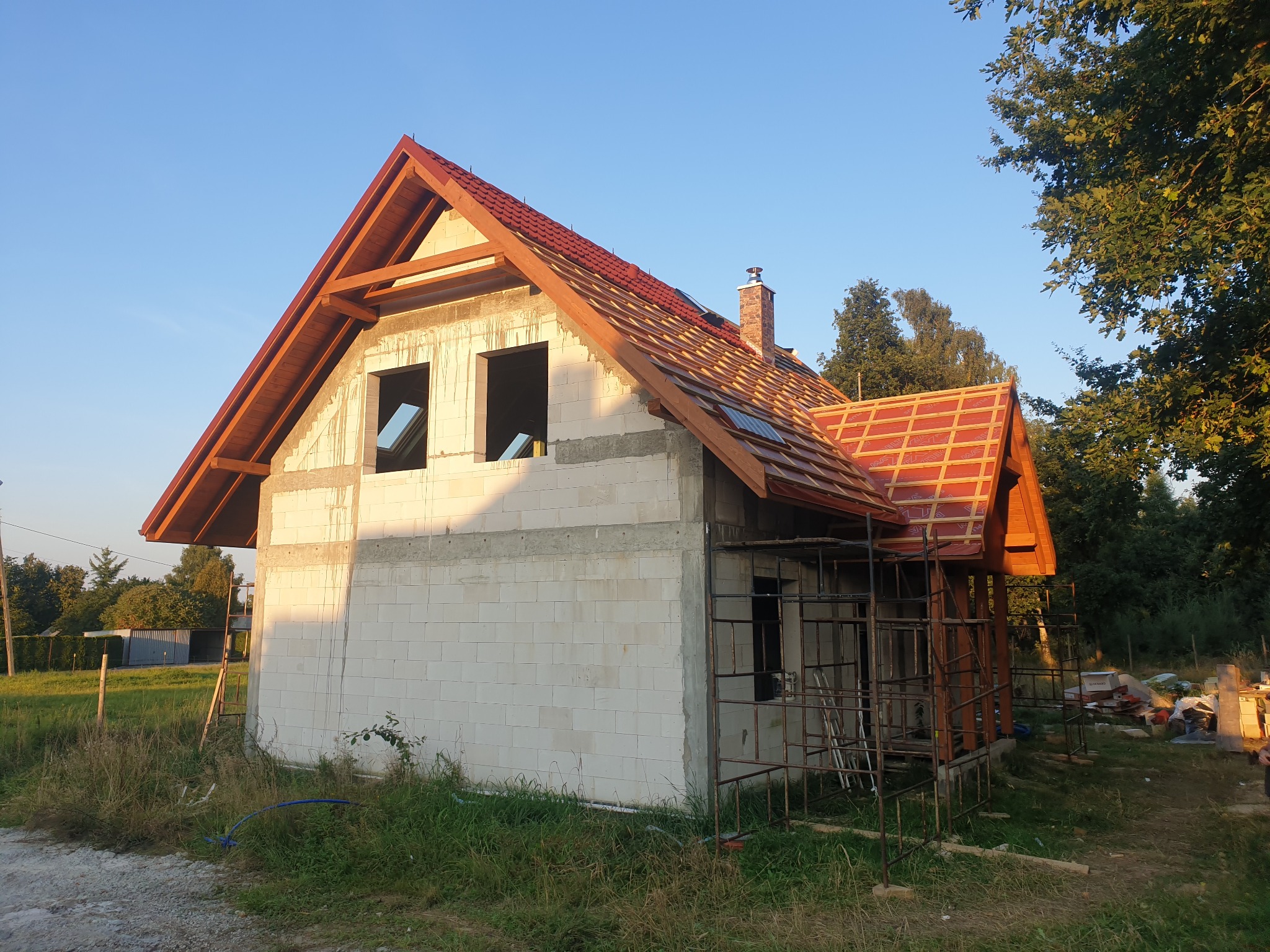 Budowa domu jednorodzinnego w Mikołowie: ściany z białych bloczków, częściowo pokryty dach z czerwonej dachówki, widoczne rusztowania i komin.