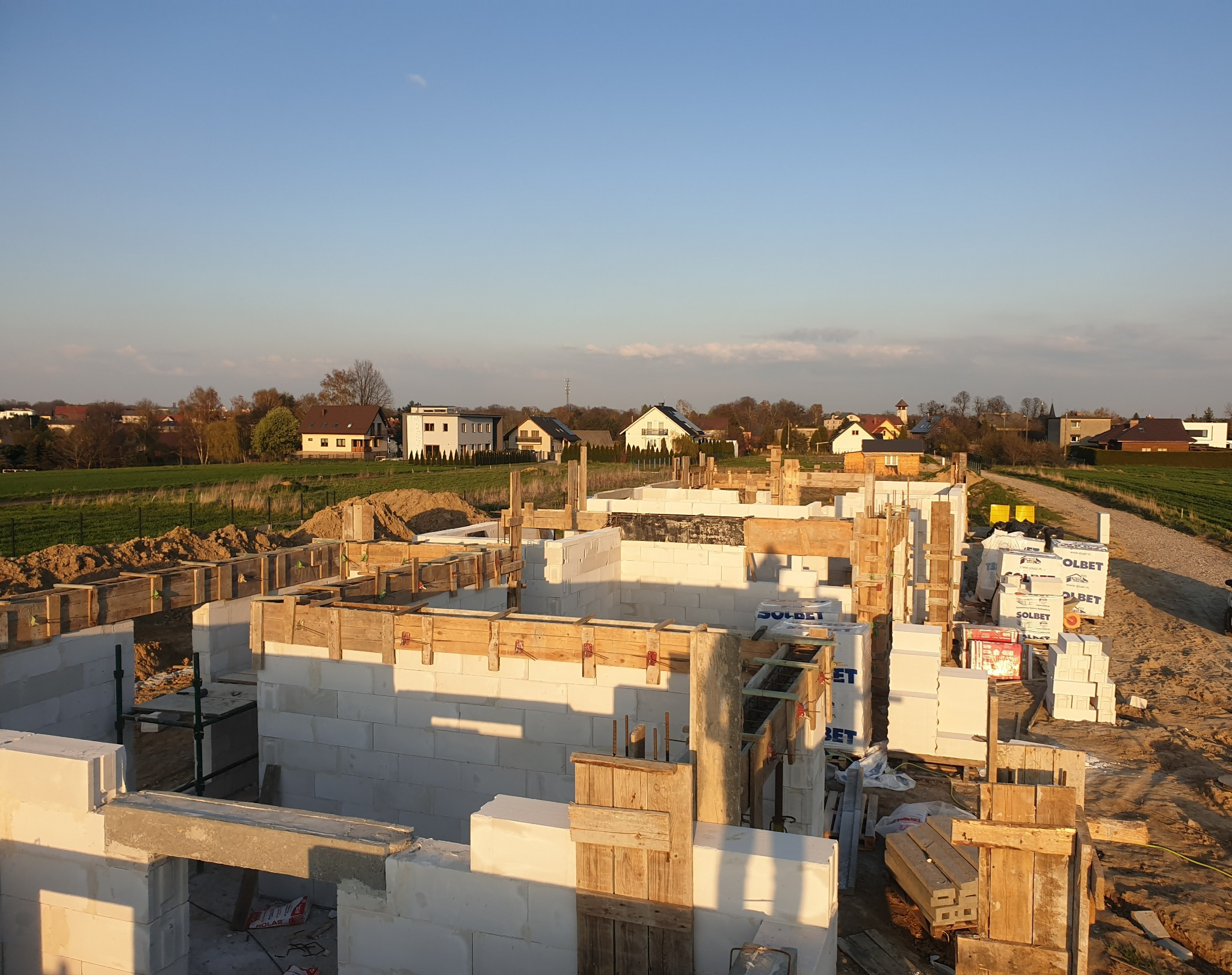 Budowa domu jednorodzinnego w Żernicy koło Gliwic: fundamenty z bloczków betonowych, szalunki drewniane, widok na okoliczne domy i pola w ciepłym świetle zachodzącego słońca.