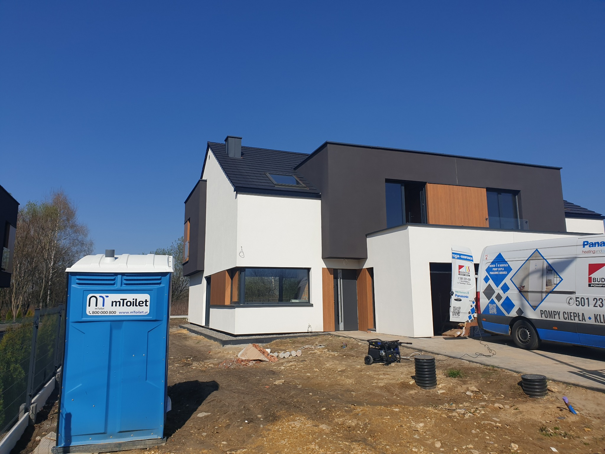 Nowoczesny dom w trakcie budowy w Czeladzi, widoczna przenośna toaleta, agregat prądotwórczy i samochód dostawczy z reklamą pomp ciepła.