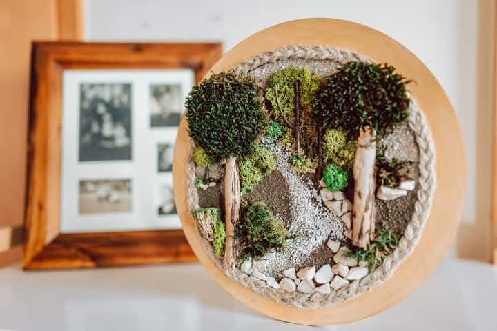 Okrągła dekoracja ścienna z miniaturowym lasem, z drzewkami z mchu i ścieżką z drobnych kamyczków, w tle drewniana ramka ze zdjęciami.