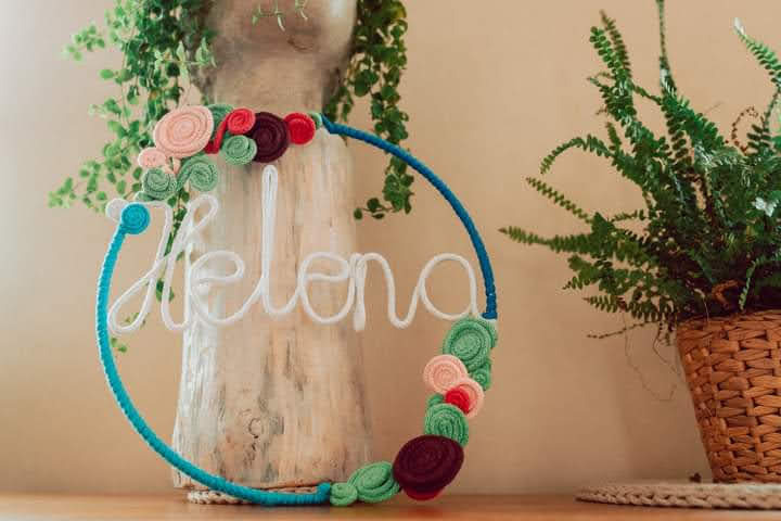 Ręcznie robiona dekoracja w kształcie koła z napisem 'Helena' wykonanym z białego sznurka, ozdobiona kolorowymi, dzierganymi kwiatami, na tle drewnianej dekoracji i paprotki w wiklinowym koszu.