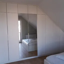 Biała szafa wnękowa z lustrzanymi drzwiami, dopasowana do skosu poddasza, widoczne łóżko w odbiciu lustra.