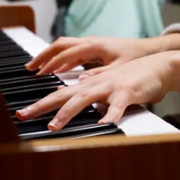 Dłonie grające na pianinie, ujęcie z bliska, widoczne klawisze i fragment drewnianej obudowy instrumentu.