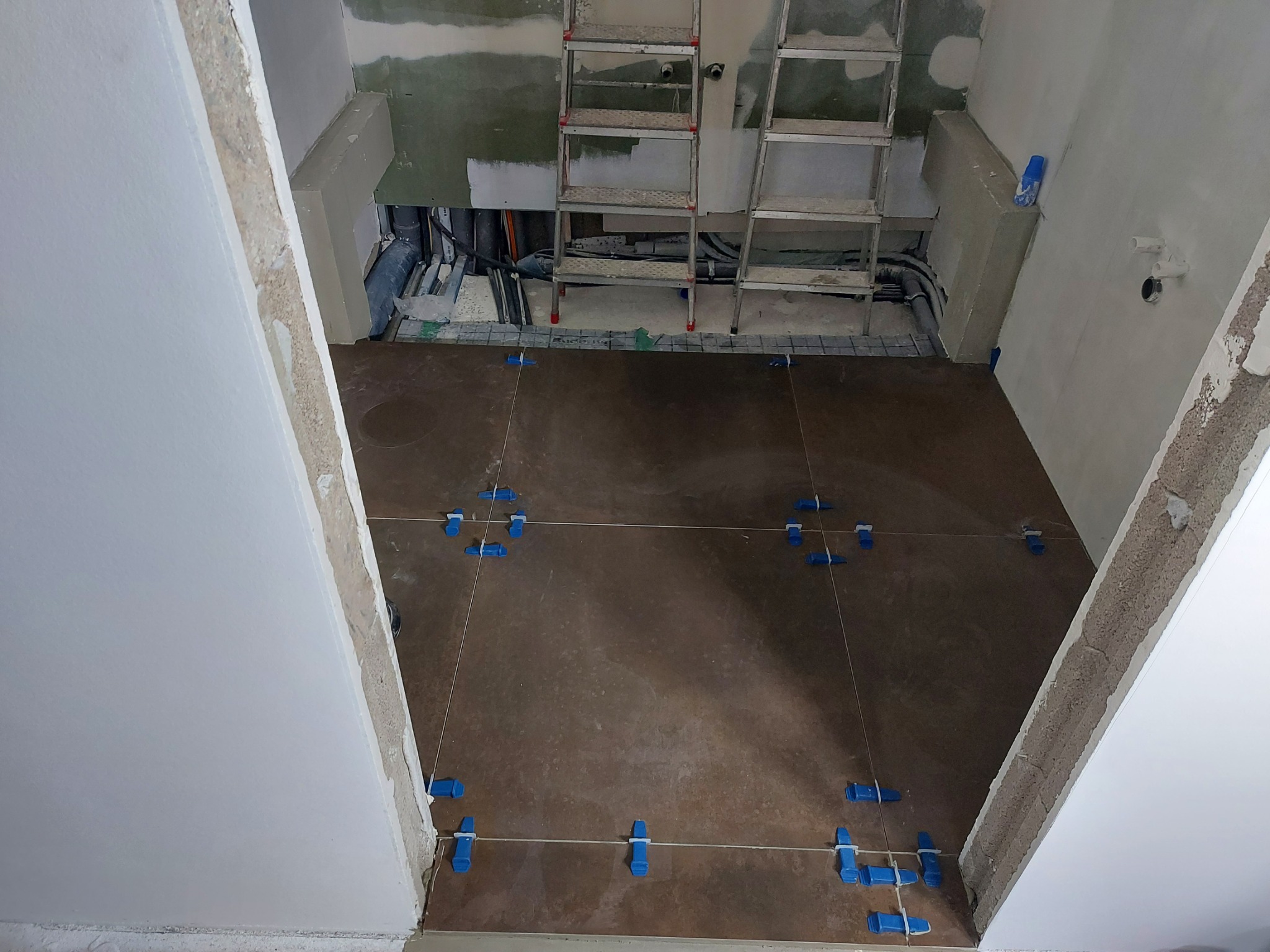 Układanie brązowych płytek podłogowych z użyciem niebieskich krzyżyków dystansowych, widoczne instalacje hydrauliczne i drabina w tle, surowy stan ścian.