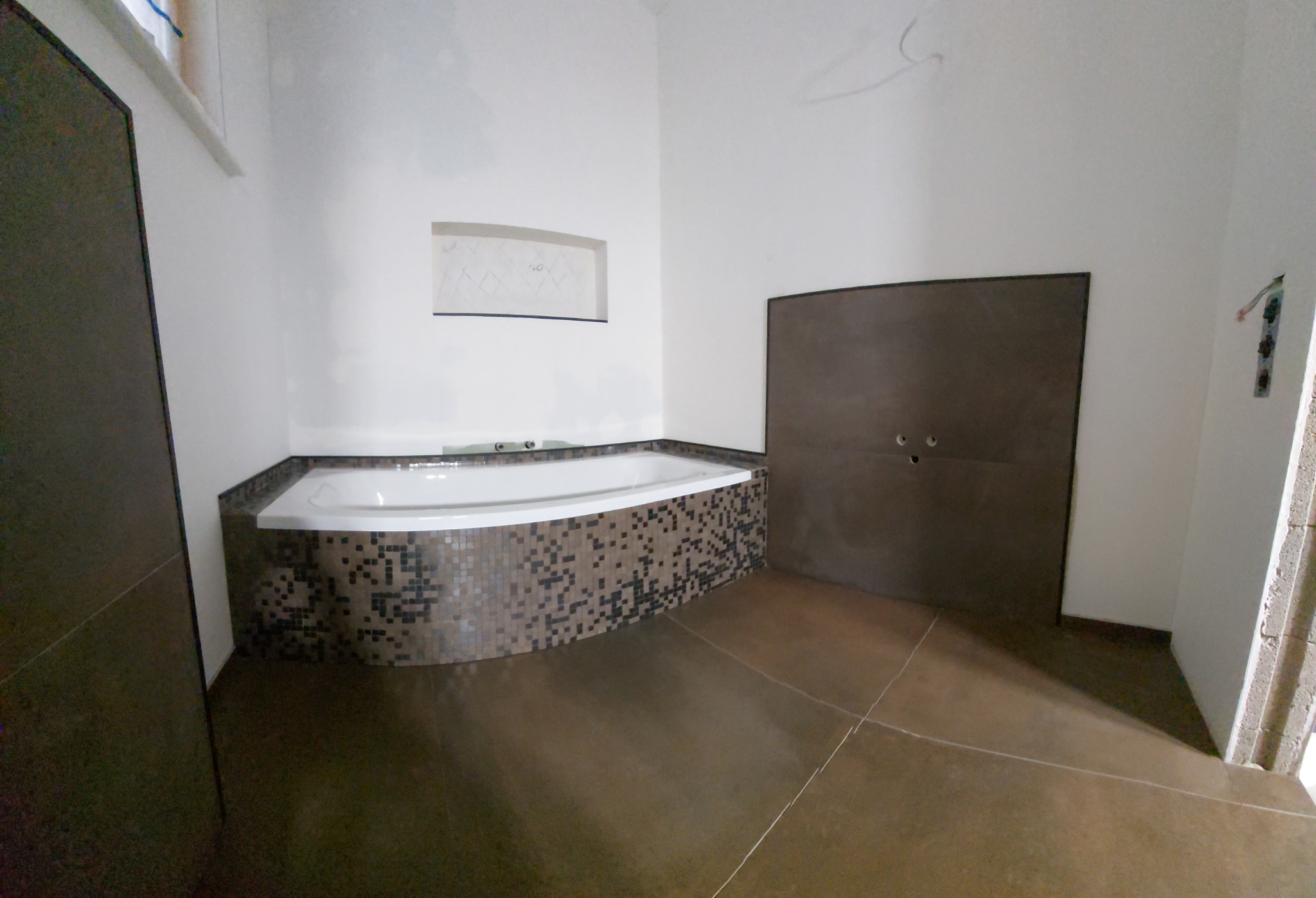 Niedokończona łazienka z wanną obudowaną mozaiką w odcieniach brązu i szarości, brązowa podłoga z widocznymi fugami, ściany w trakcie wykończenia, widoczna instalacja elektryczna bez osprzętu.