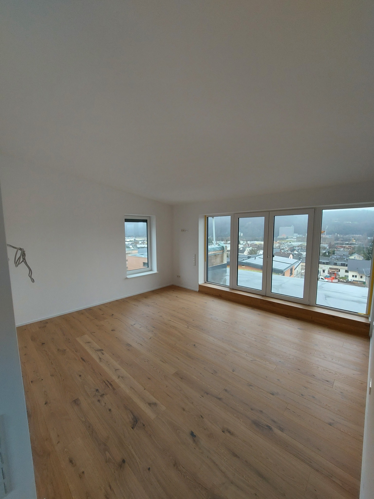 Puste, wykończone wnętrze z drewnianą podłogą, białymi ścianami i dużymi oknami z widokiem na miasto w tle, widoczne wystające kable elektryczne.