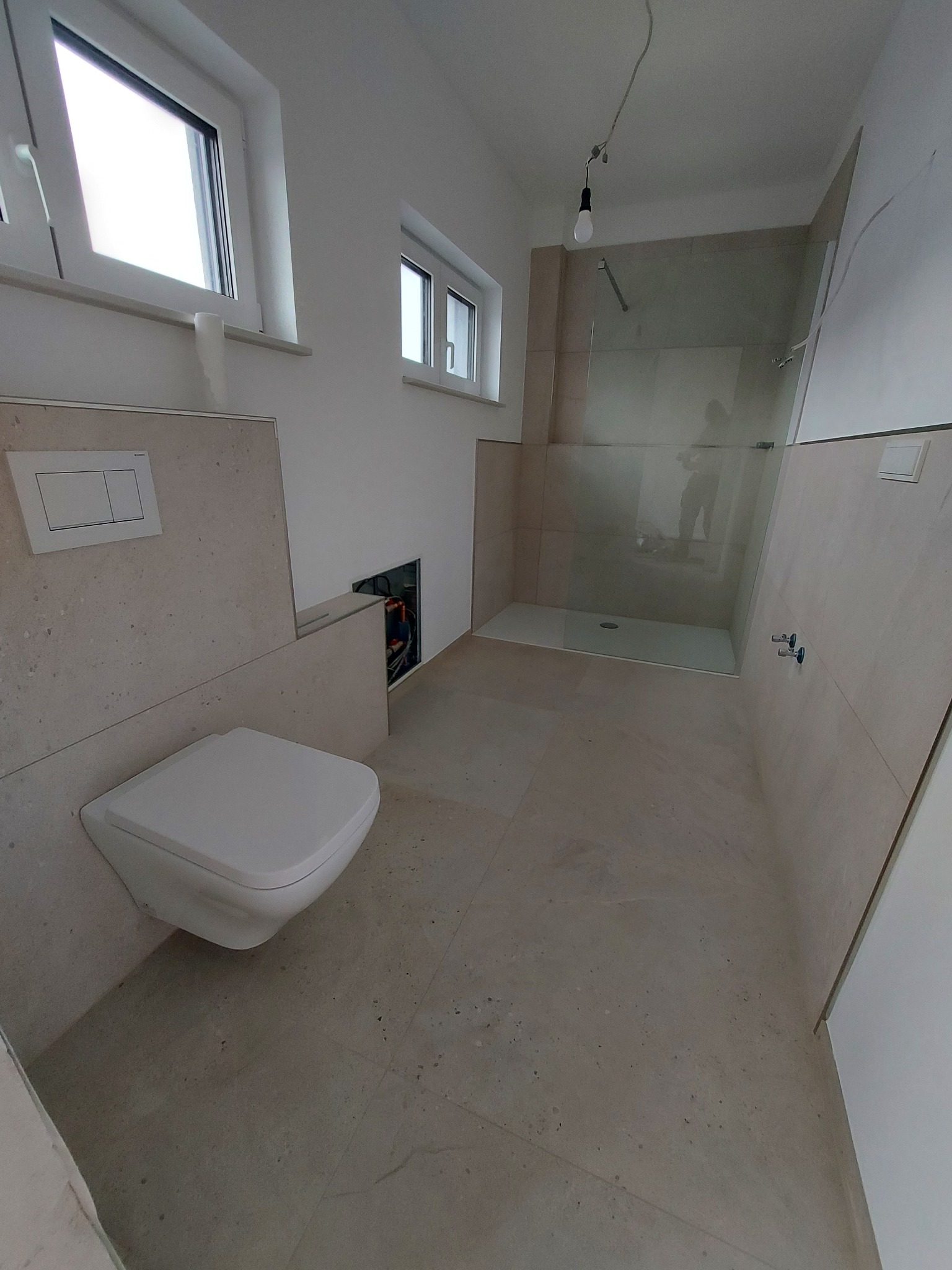 Nowoczesna łazienka w trakcie wykończenia: podwieszana toaleta, kabina prysznicowa ze szklaną ścianą, beżowe płytki imitujące kamień, widoczne instalacje w ścianie.