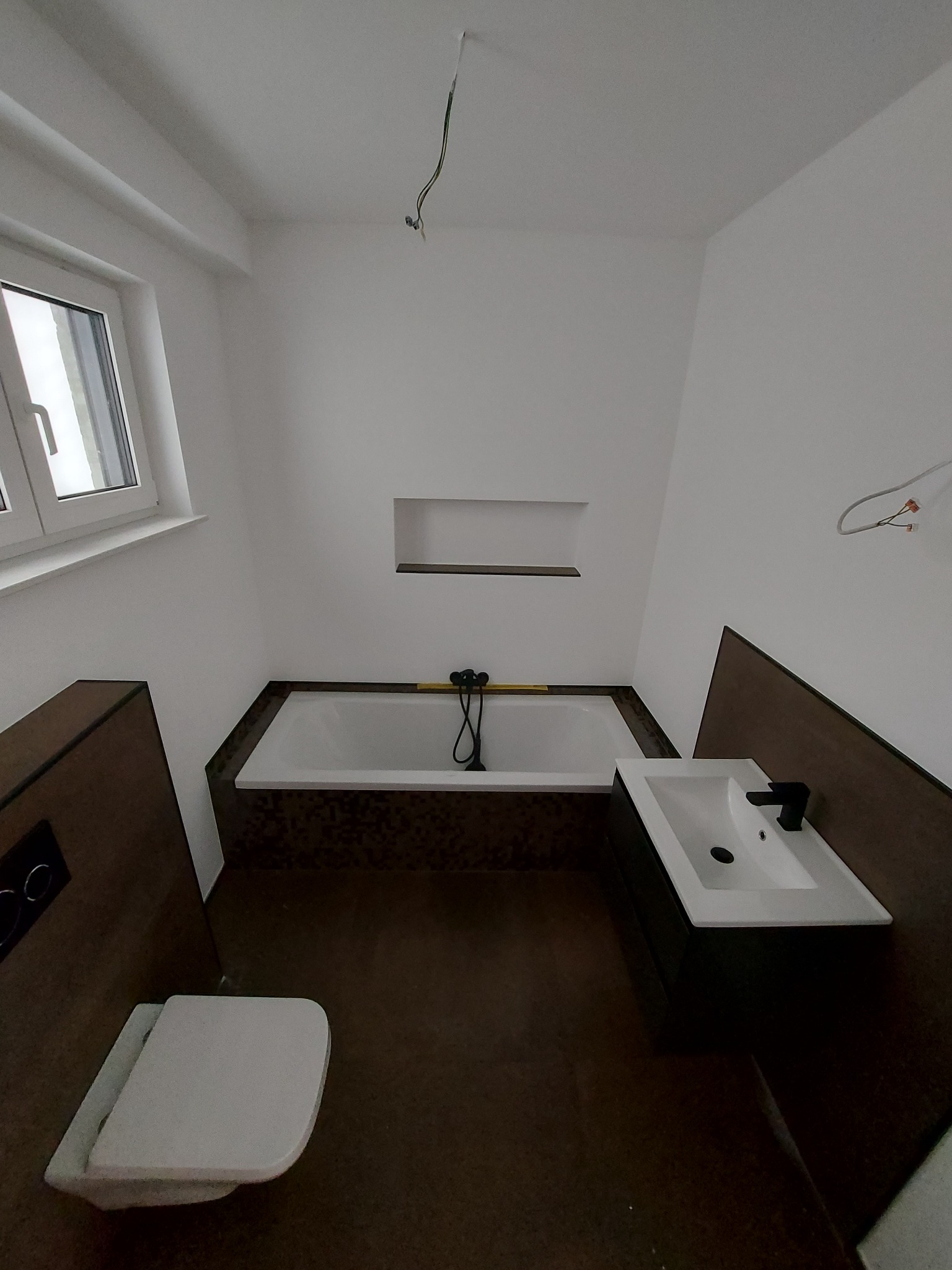 Wnętrze łazienki w trakcie prac wykończeniowych: biała wanna obudowana brązowymi płytkami, umywalka z czarną armaturą, podwieszana toaleta i wystające kable z sufitu.