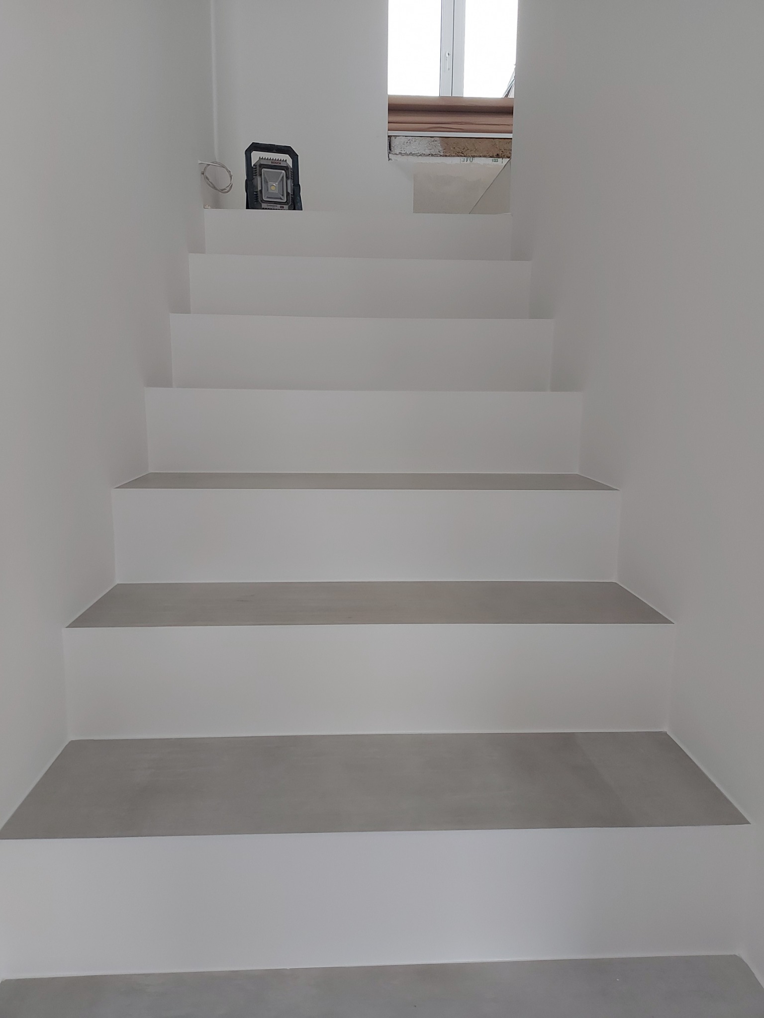 Minimalistyczne, białe schody z szarymi stopnicami w nowym domu, oświetlone lampą roboczą na górnym stopniu, widok z dołu.