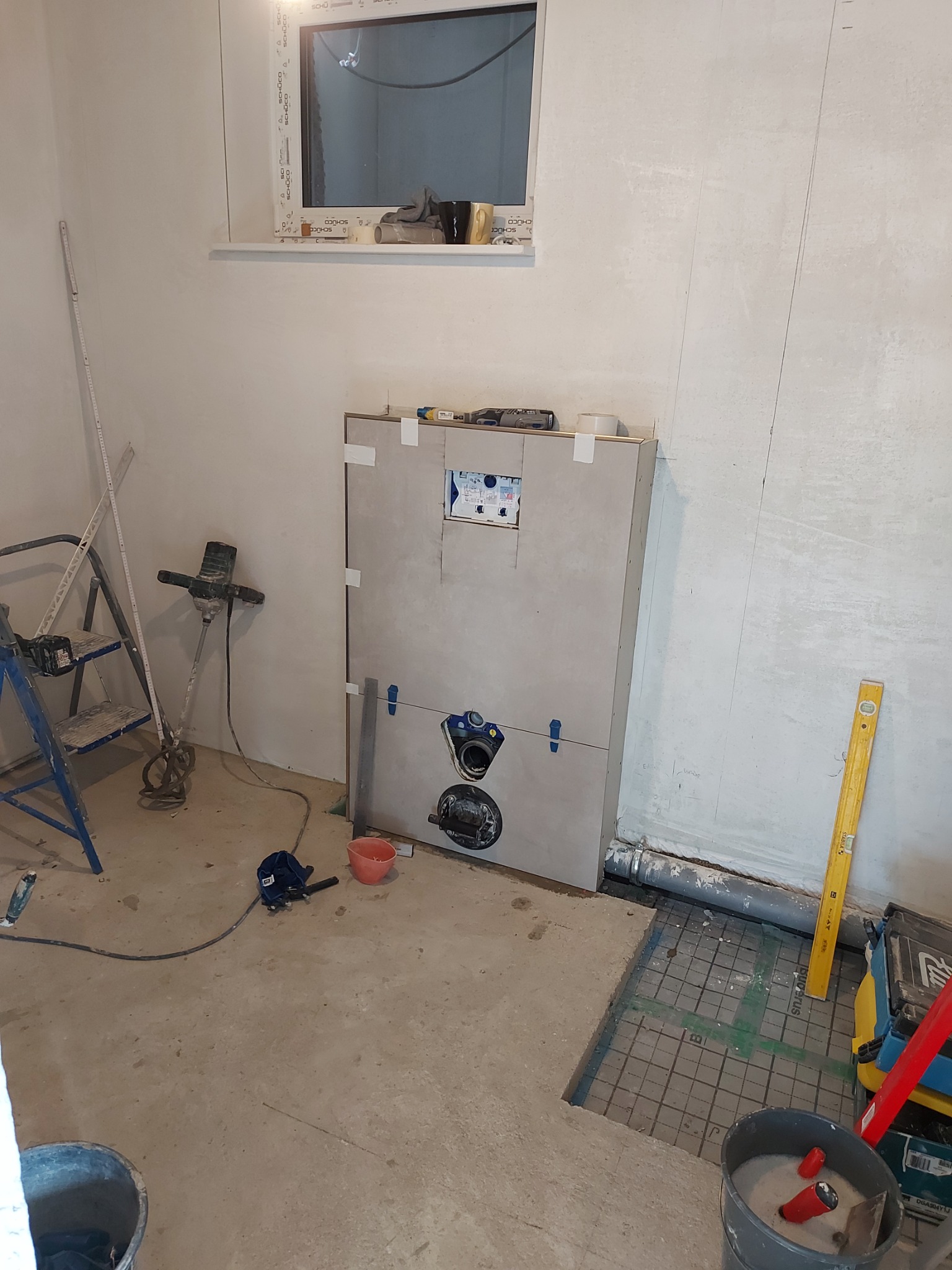 Przedścienna instalacja WC w trakcie montażu, widoczne elementy mocujące, poziomica, mieszadło do zaprawy, wiadro z zaprawą, rury kanalizacyjne i okno w surowym stanie