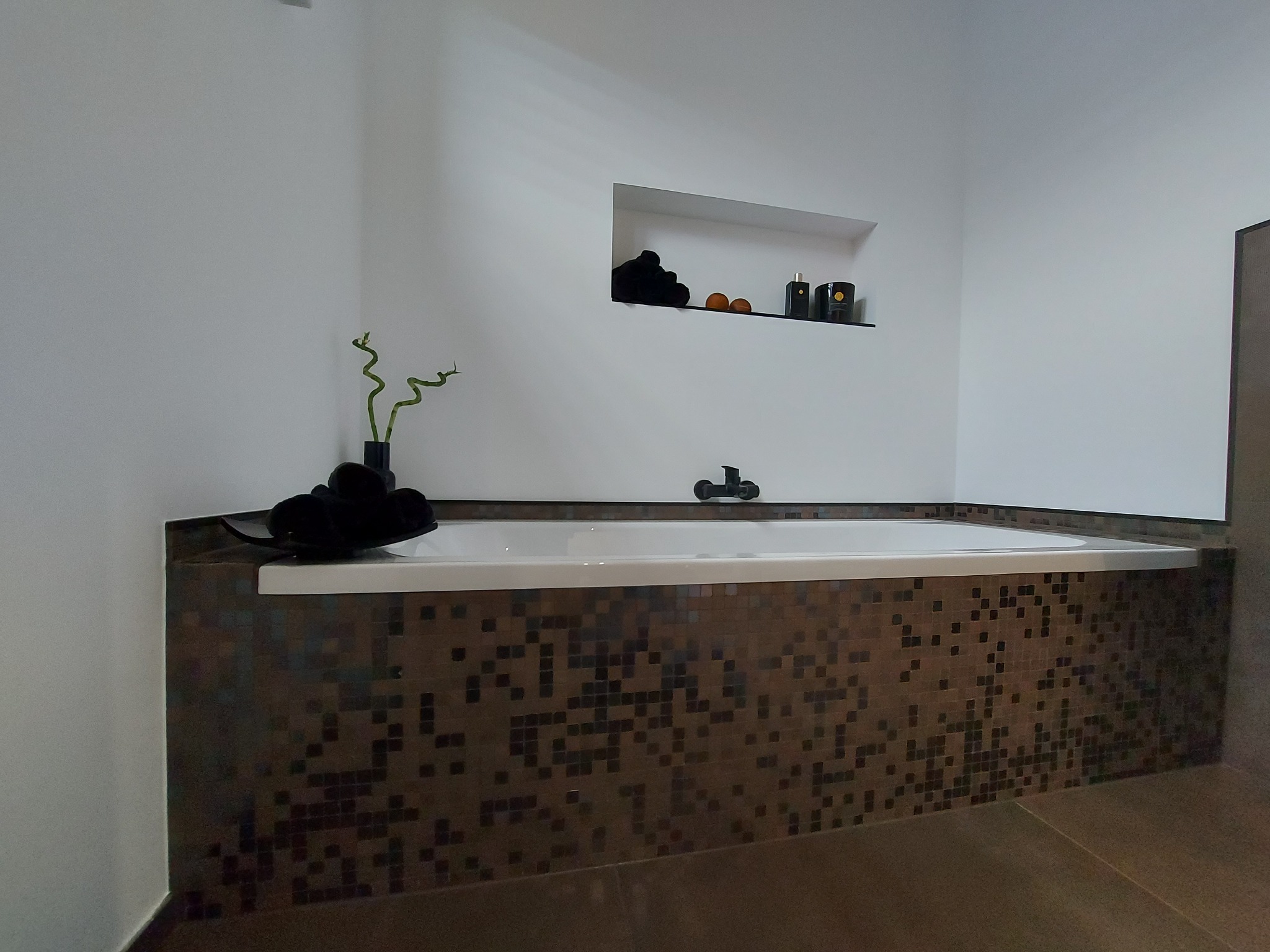 Nowoczesna łazienka z wanną obudowaną mozaiką w odcieniach brązu, na wannie czarne ręczniki, w ścianie wnęka z dekoracjami i oświetleniem punktowym, bambus w czarnym wazonie.