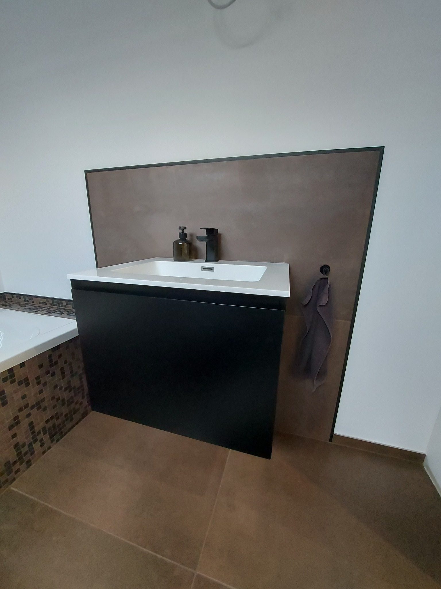 Nowoczesna łazienka z czarną szafką pod umywalką, białą umywalką, brązowym panelem ściennym i dopasowanymi brązowymi płytkami podłogowymi oraz dozownikiem mydła i czarną baterią.