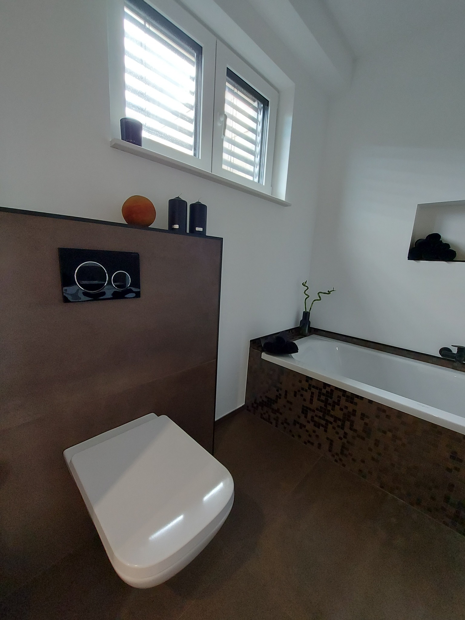 Nowoczesna łazienka z podwieszaną toaletą, brązowymi mozaikami na obudowie wanny i oknem z żaluzjami.