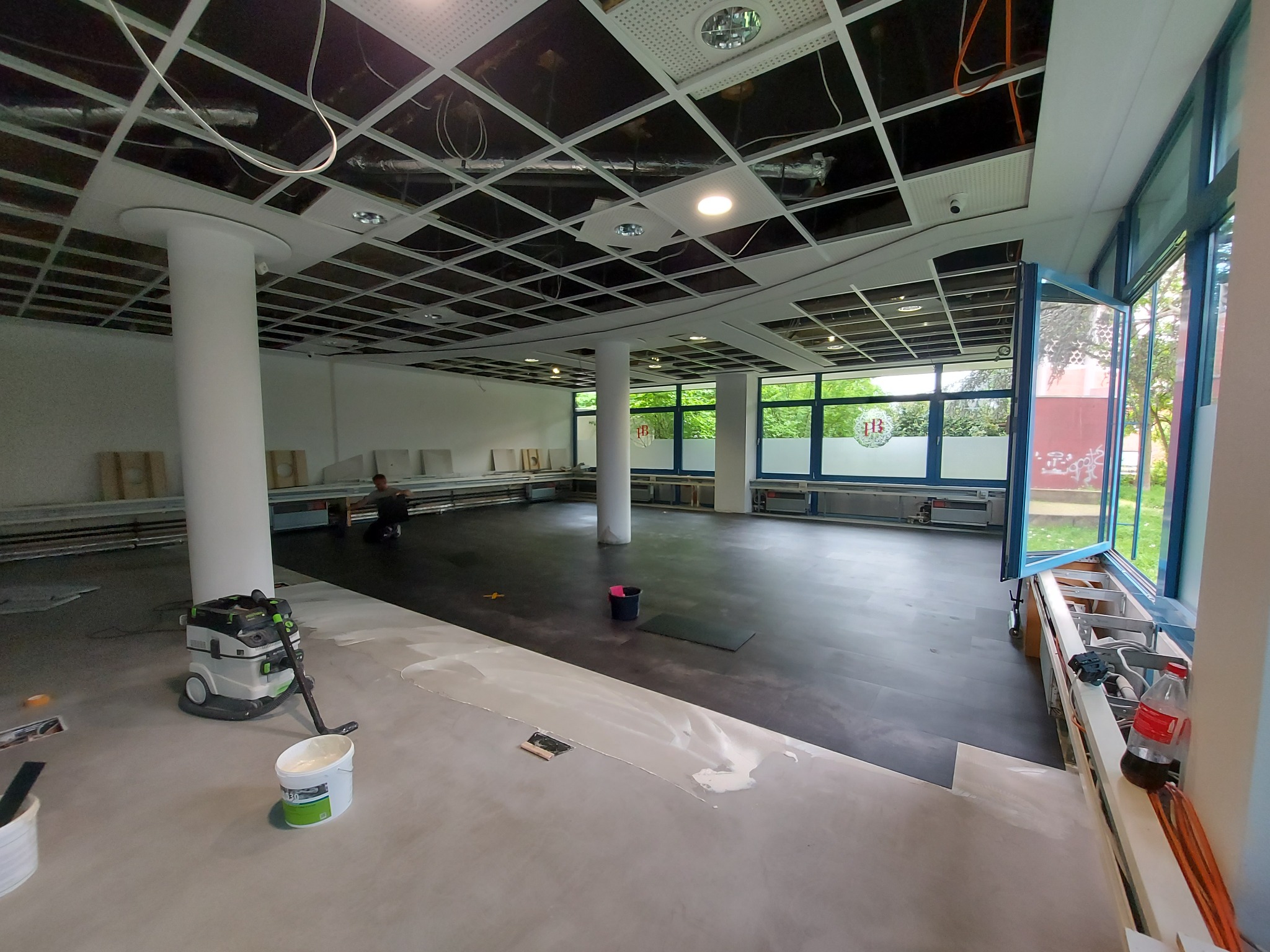 Układanie ciemnej wykładziny w przestronnym pomieszczeniu biurowym z kolumnami, widoczny odkurzacz przemysłowy, wiadro z klejem i częściowo odsłonięty sufit podwieszany.