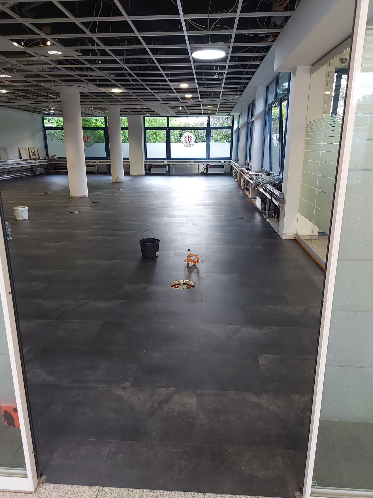 Wnętrze biura w trakcie remontu: czarna podłoga w płytkach, sufit z odsłoniętą konstrukcją, białe filary i okna z widokiem na zieleń, widoczne narzędzia pracy i wiadro.