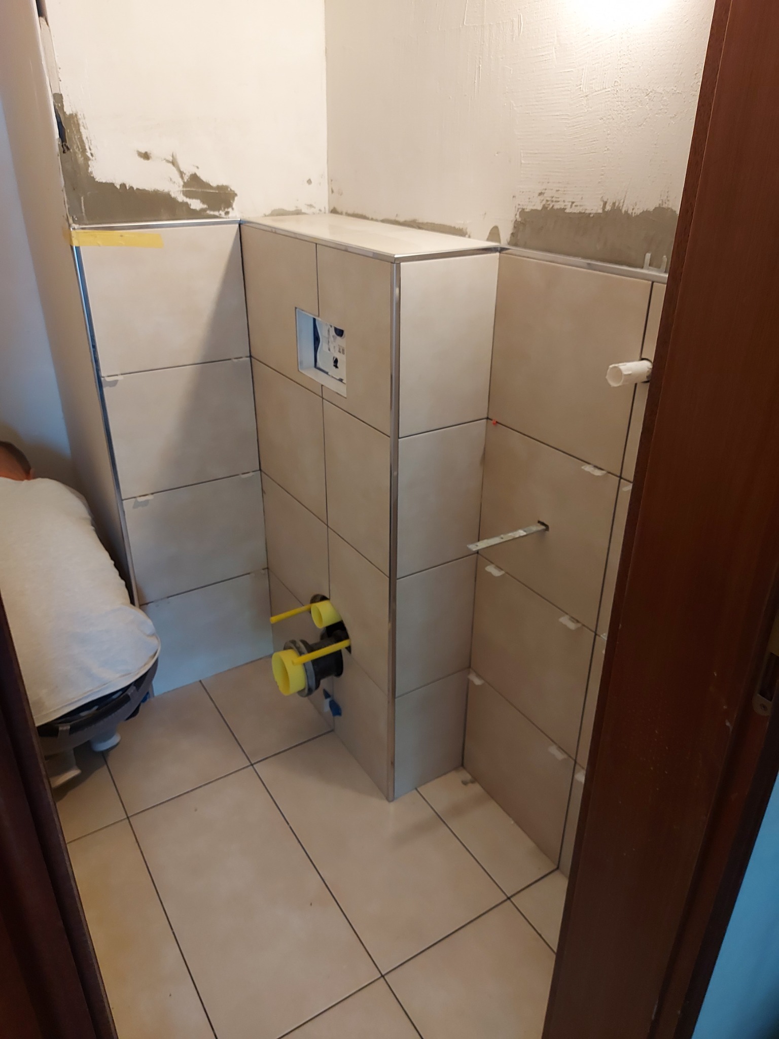 Montaż płytek w łazience, widoczne fragmenty instalacji wodnej i kanalizacyjnej, płytki beżowe z fugami, częściowo wykończone ściany, widoczne listwy wykończeniowe i poziomica.