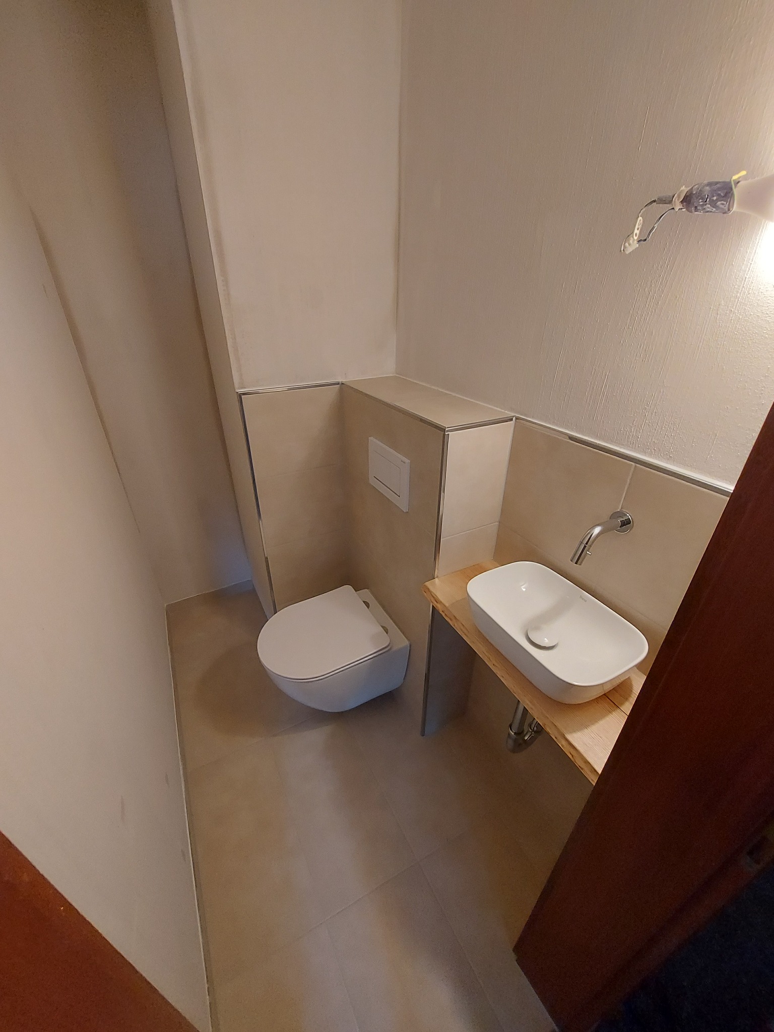 Nowoczesna toaleta z podwieszaną miską WC i umywalką nablatową na drewnianym blacie, jasne płytki ścienne, widoczne niedokończone prace elektryczne