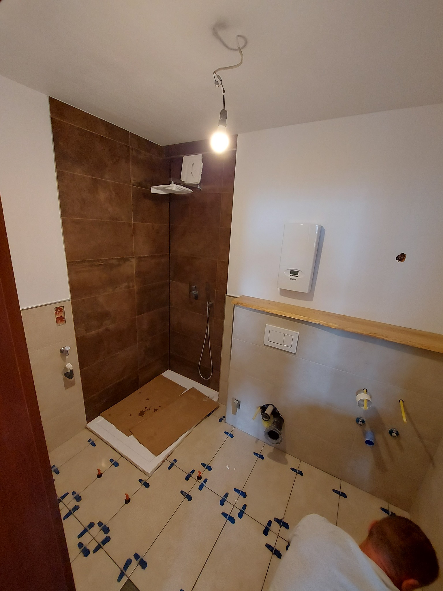 Łazienka w trakcie remontu: brązowe płytki w kabinie prysznicowej, jasne płytki podłogowe z niebieskimi krzyżykami dystansowymi, widoczne rury i instalacje przed zabudową.