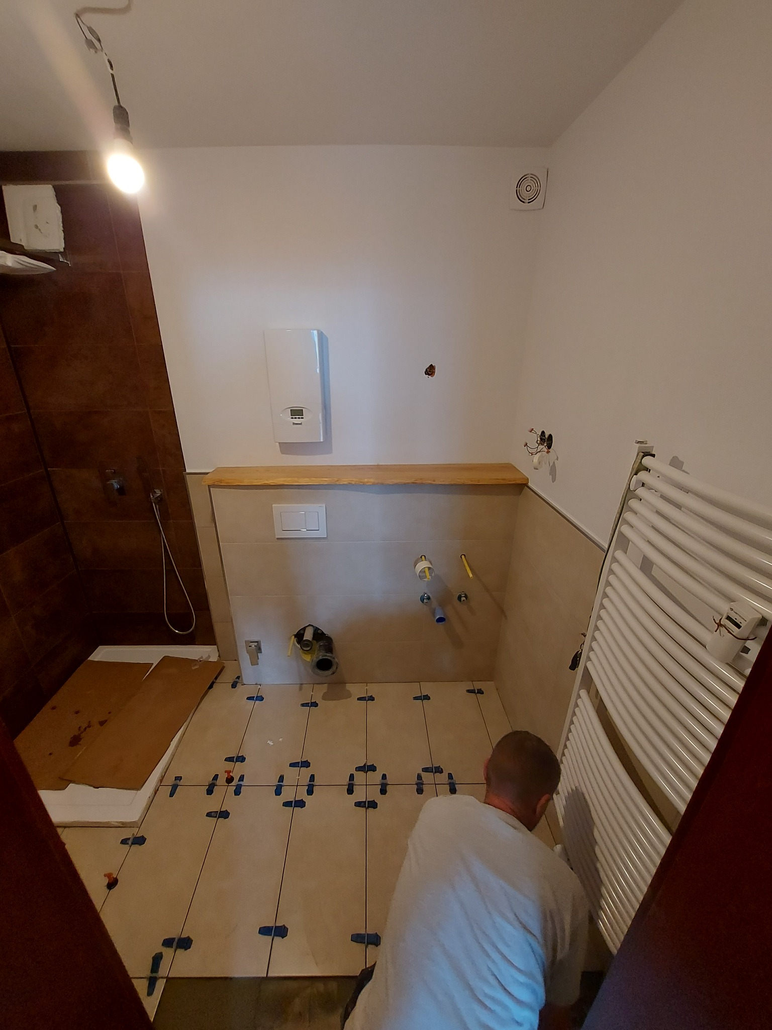 Łazienka w trakcie układania płytek podłogowych z użyciem klipsów poziomujących, widoczny grzejnik drabinkowy, przygotowane przyłącza wodno-kanalizacyjne i częściowo wykończona ściana z drewnianym...