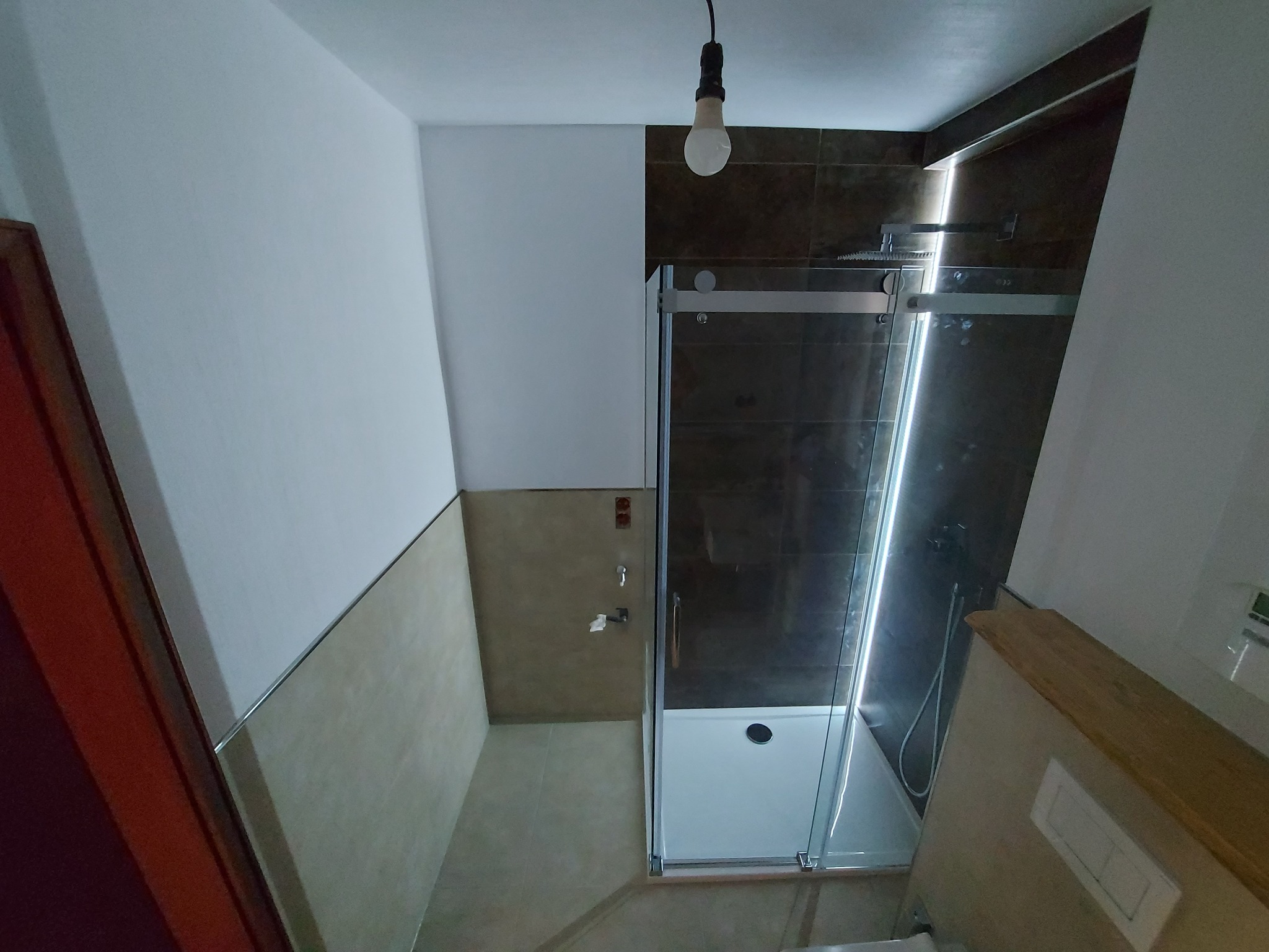 Wnętrze łazienki po remoncie, widoczna kabina prysznicowa z przesuwnymi drzwiami, brązowe płytki na ścianie pod prysznicem, beżowe płytki na pozostałej części ściany, surowa żarówka zwisająca...