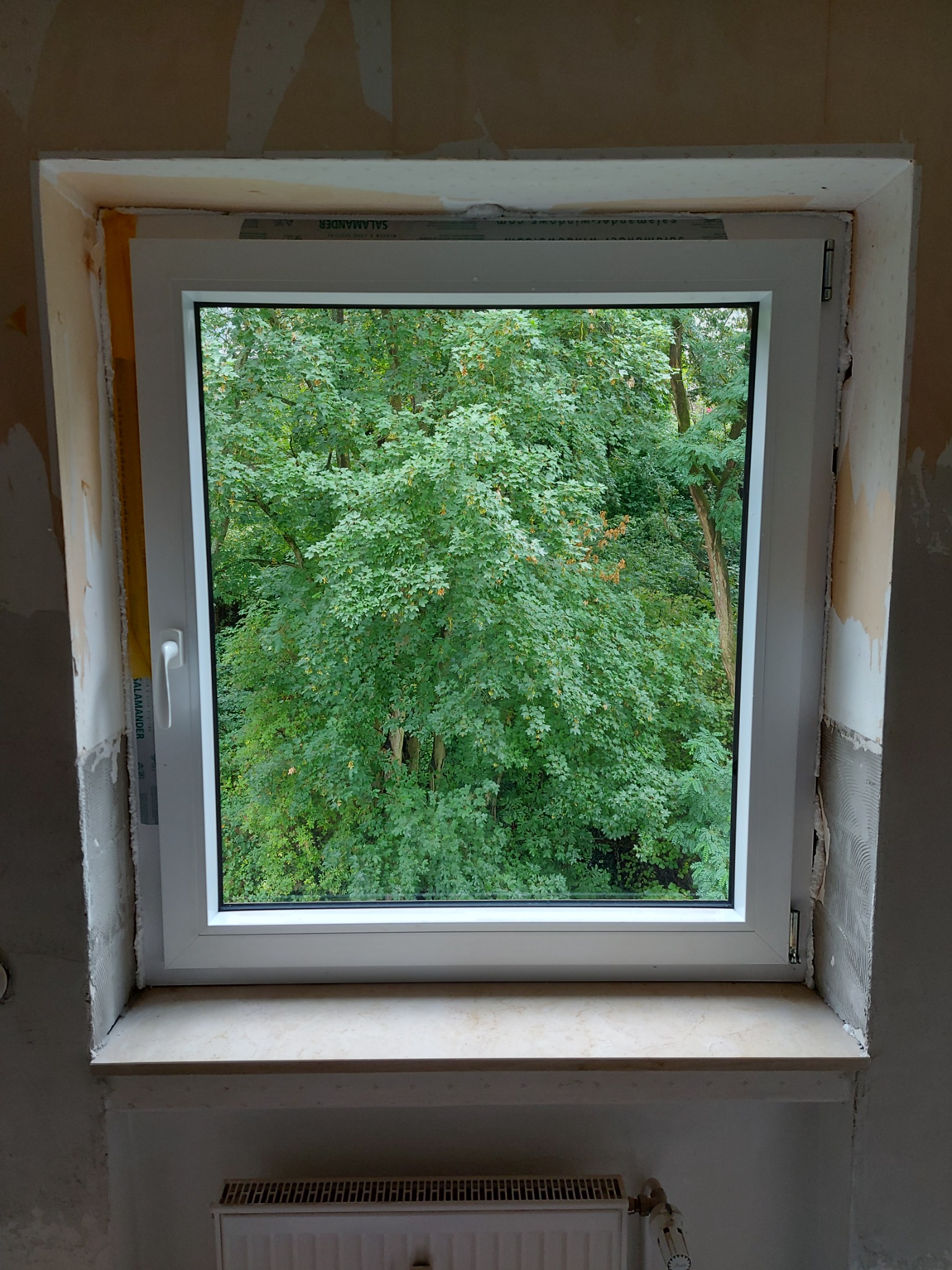 Nowe, białe okno PCV zamontowane w surowym otworze okiennym, widoczne ślady tynku i niedokończone wykończenie wokół ramy, na zewnątrz bujna zieleń drzew.