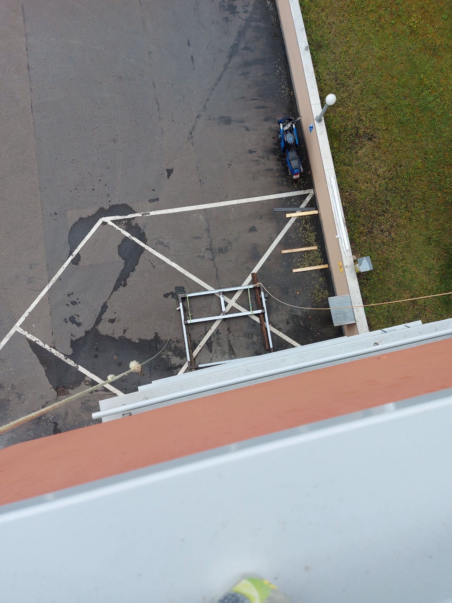 Widok z góry na parking z metalową konstrukcją, motocyklem i plamami na asfalcie wewnątrz białych linii wyznaczających miejsce postojowe, obok budynek z trawnikiem.