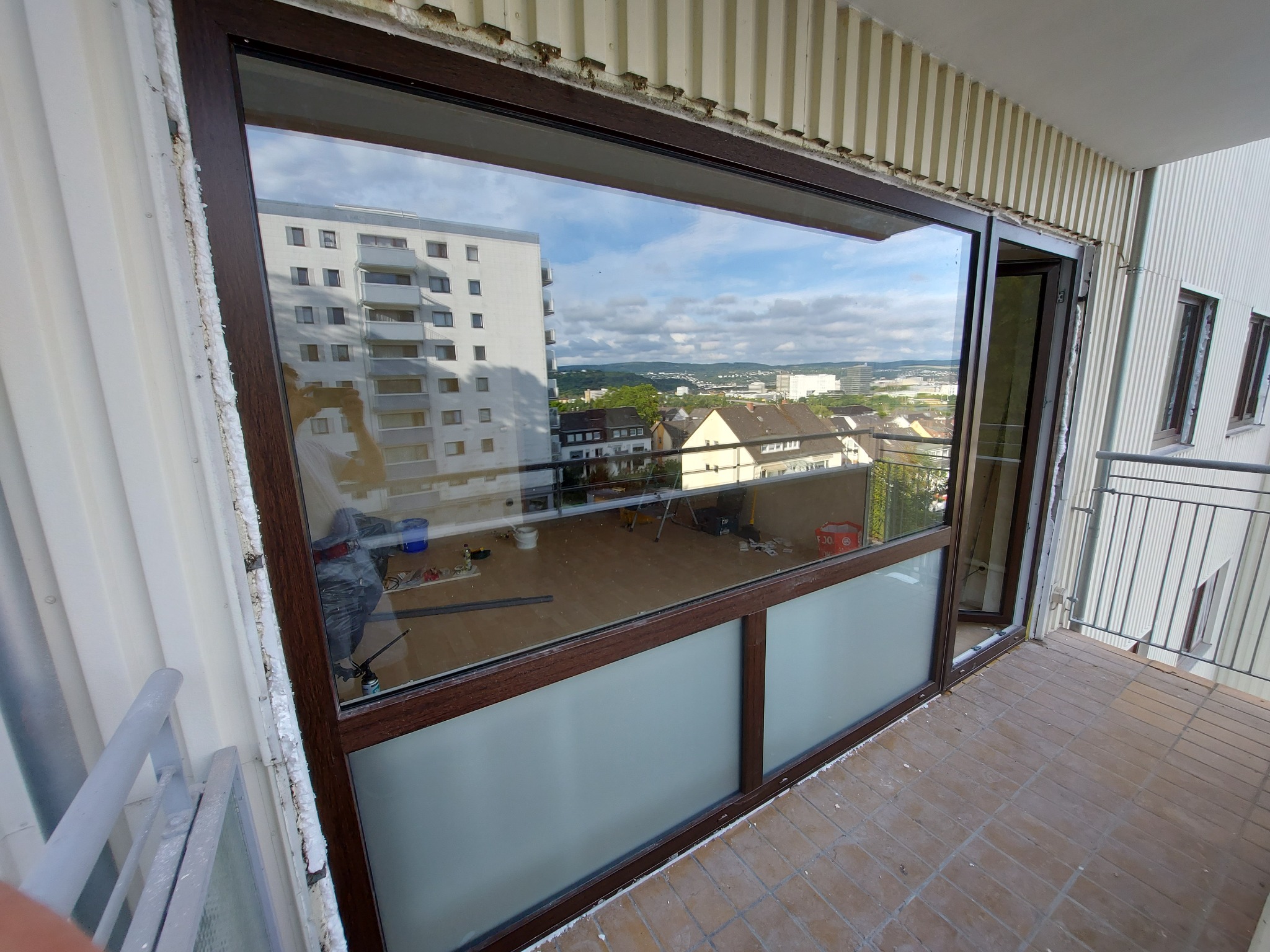 Montaż okna balkonowego z brązową ramą, widoczne pianowanie uszczelniające, na podłodze balkonowej brązowe płytki, w odbiciu okna widoczny budynek mieszkalny i krajobraz miejski.