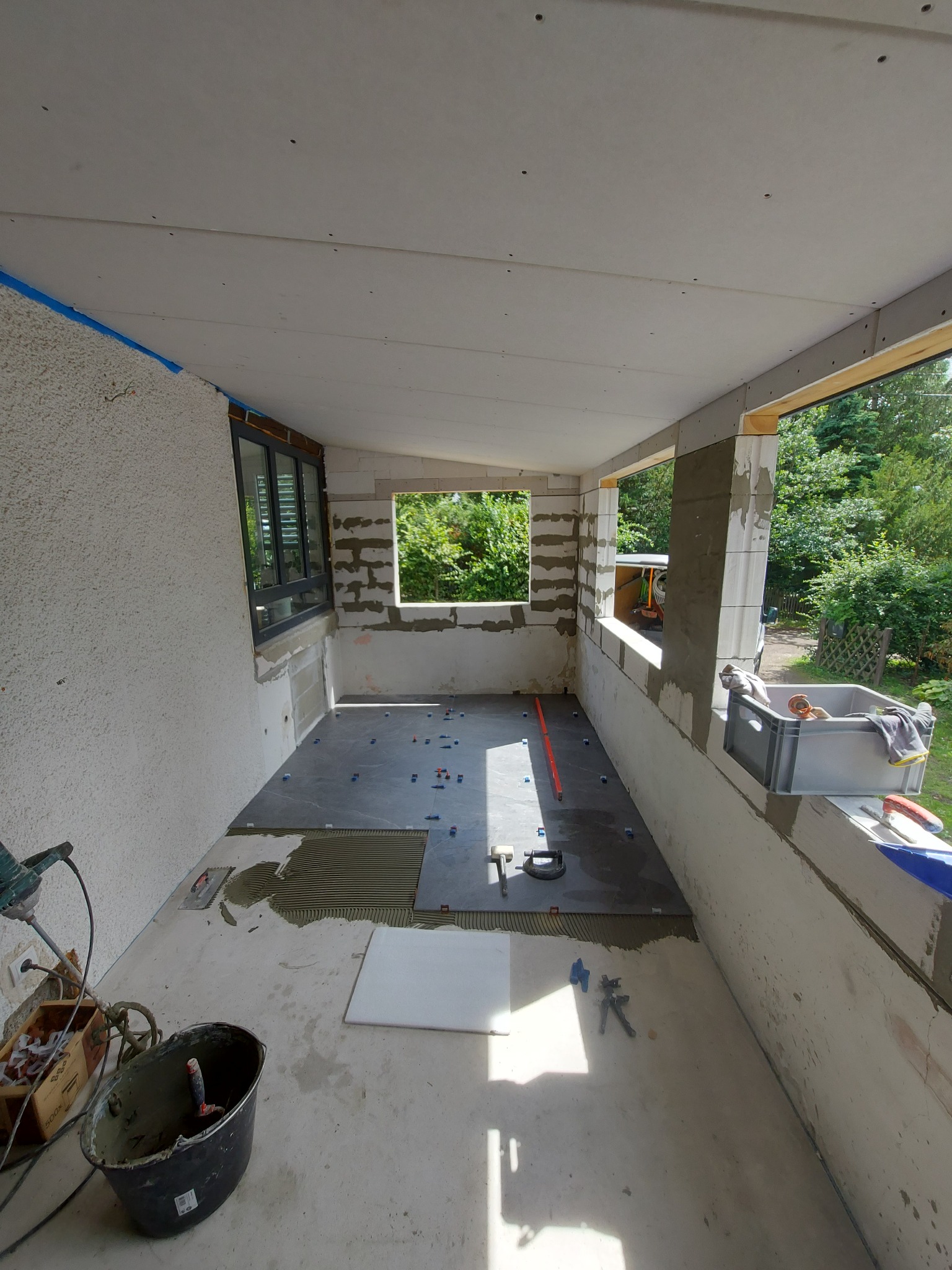 Montaż ciemnoszarych płytek podłogowych na balkonie w trakcie remontu, widoczne narzędzia, poziomica i krzyżyki dystansowe, ściany w stanie surowym, widok na zieleń za oknem.