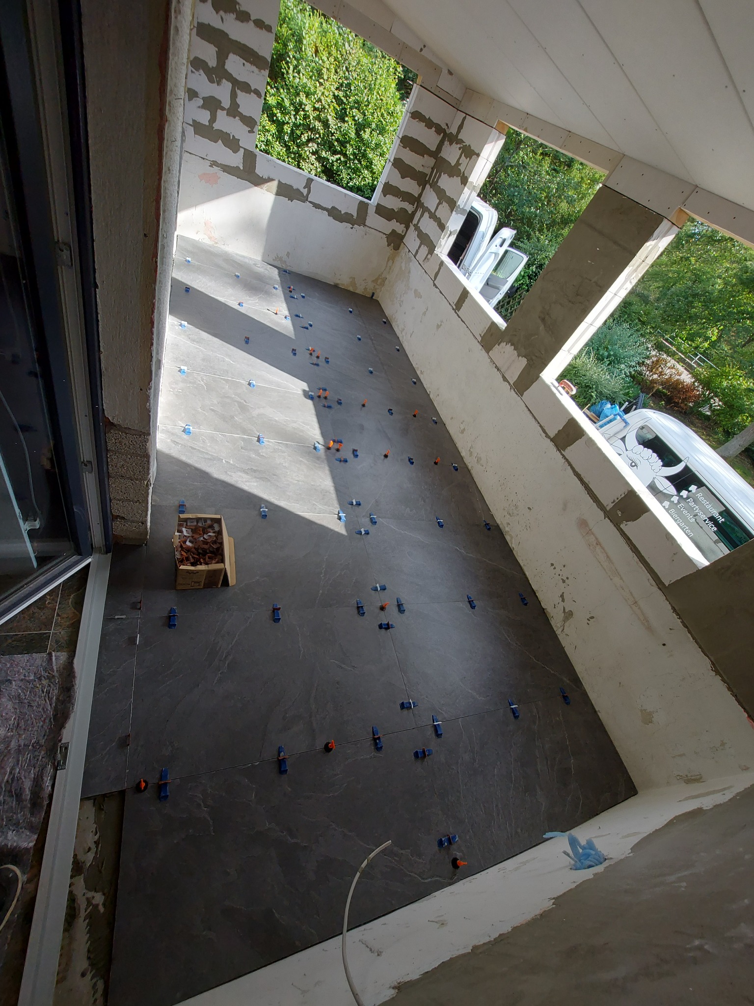 Układanie ciemnoszarych płytek podłogowych na balkonie, widoczne plastikowe krzyżyki dystansowe i karton z klinami poziomującymi, surowe ściany w tle.