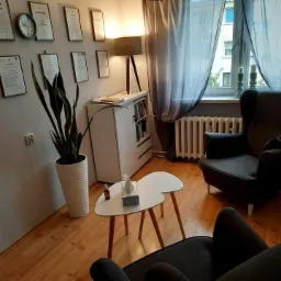 Jasne wnętrze gabinetu z dwoma ciemnymi fotelami, stolikiem w kształcie nerki, regałem z książkami i ścianą udekorowaną certyfikatami