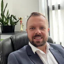 Portret mężczyzny w garniturze siedzącego na skórzanym fotelu biurowym, z rośliną doniczkową i trofeami w tle.