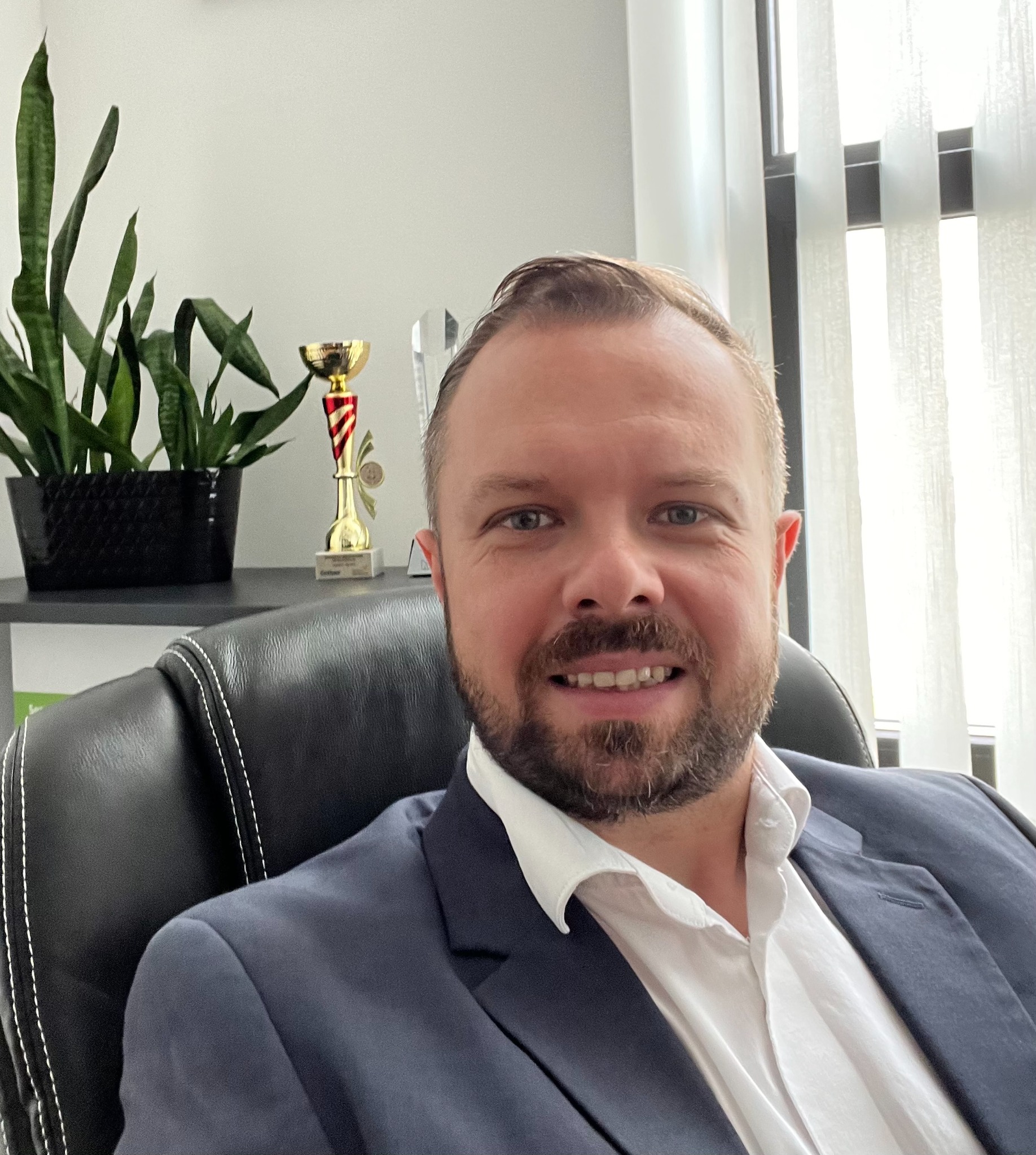 Portret mężczyzny w garniturze siedzącego na skórzanym fotelu biurowym, z rośliną doniczkową i trofeami w tle.