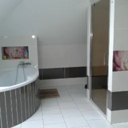 Biała łazienka na poddaszu z wanną obudowaną szarymi płytkami w paski, kabiną prysznicową i dekoracyjnymi panelami z motywem kwiatowym.