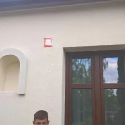 Elewacja budynku z oknem, wnęką w kształcie łuku i fragmentem zabezpieczenia na ścianie przed malowaniem, widoczna faktura tynku.