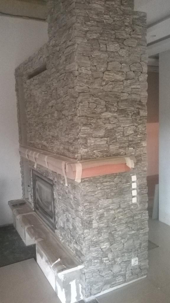 Obudowa kominka wykończona szarą, nieregularną okładziną kamienną, zabezpieczona folią i taśmą malarską podczas prac wykończeniowych.